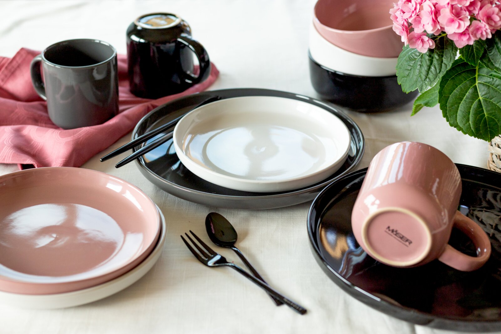 Modernes Keramik-Geschirrset in Schwarz, Weiß und Rosé mit Tellern, Schalen, Tassen und Besteck auf gedecktem Tisch, fotografiert aus leicht erhöhter schräger Perspektive (3/4-Ansicht)