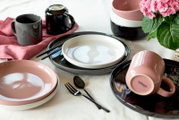 Modernes Keramik-Geschirrset in Schwarz, Weiß und Rosé mit Tellern, Schalen, Tassen und Besteck auf gedecktem Tisch, fotografiert aus leicht erhöhter schräger Perspektive (3/4-Ansicht)