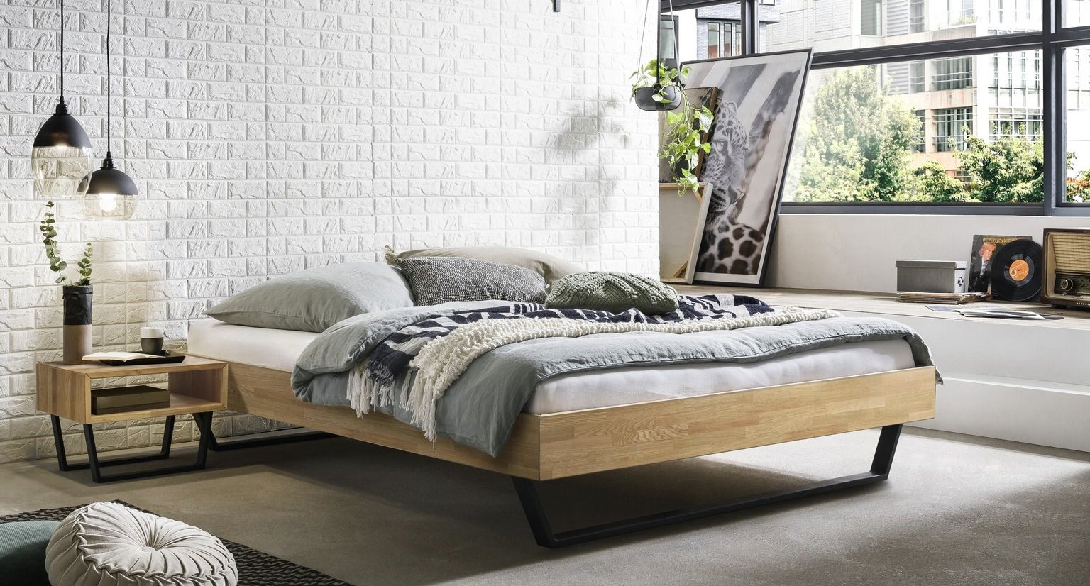 Modernes niedriges Holzbett mit schwarzem Metallgestell, bezogen mit grauer Bettwäsche und Kissen in einem hellen Schlafzimmer vor weißer Ziegelwand; Perspektive: seitliche Frontansicht aus leicht erhöhter Position.