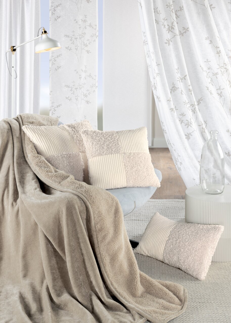 Gemütliche Wohnzimmerszene mit beigen Kissenhüllen auf einem Sofa, umgeben von einem weichen Überwurf und dekorativen Vorhängen im Hintergrund. Perspektive von vorne.