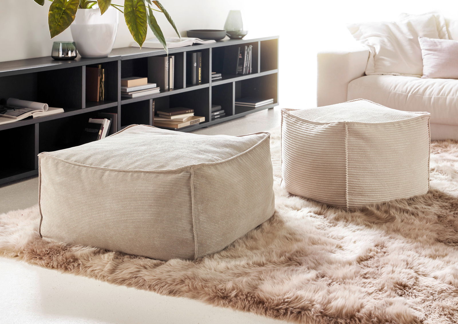 Zwei beige, quadratische Hocker aus Stoff auf einem flauschigen Teppich in einem modernen Wohnzimmer, seitliche Perspektive.