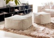 Twee beige, vierkante stoffen poefs op een zacht tapijt in een moderne woonkamer, gezien vanuit de zijkant.