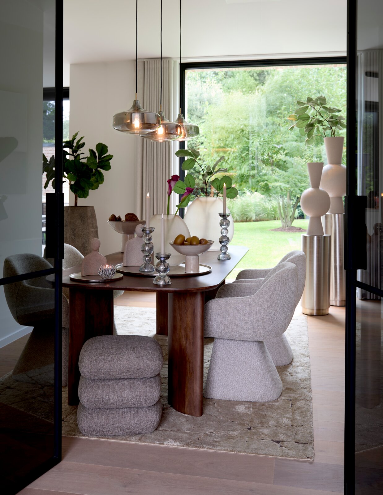 Frontalansicht durch geöffnete Glastüren auf ein elegantes Esszimmer mit massivem Holztisch, gepolsterten Designerstühlen, skulpturalen Wohnaccessoires und gläsernen Pendelleuchten vor bodentiefem Gartenfenster.