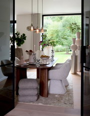 Frontalansicht durch geöffnete Glastüren auf ein elegantes Esszimmer mit massivem Holztisch, gepolsterten Designerstühlen, skulpturalen Wohnaccessoires und gläsernen Pendelleuchten vor bodentiefem Gartenfenster.