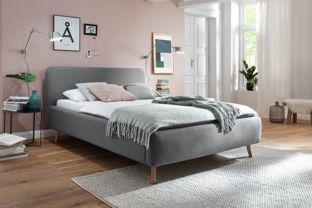 Z2 Polsterbett MATTIS Graues Polsterbett in einem modernen Schlafzimmer mit Holzfußboden, dekoriert mit Kissen und Decken, seitliche Perspektive.
