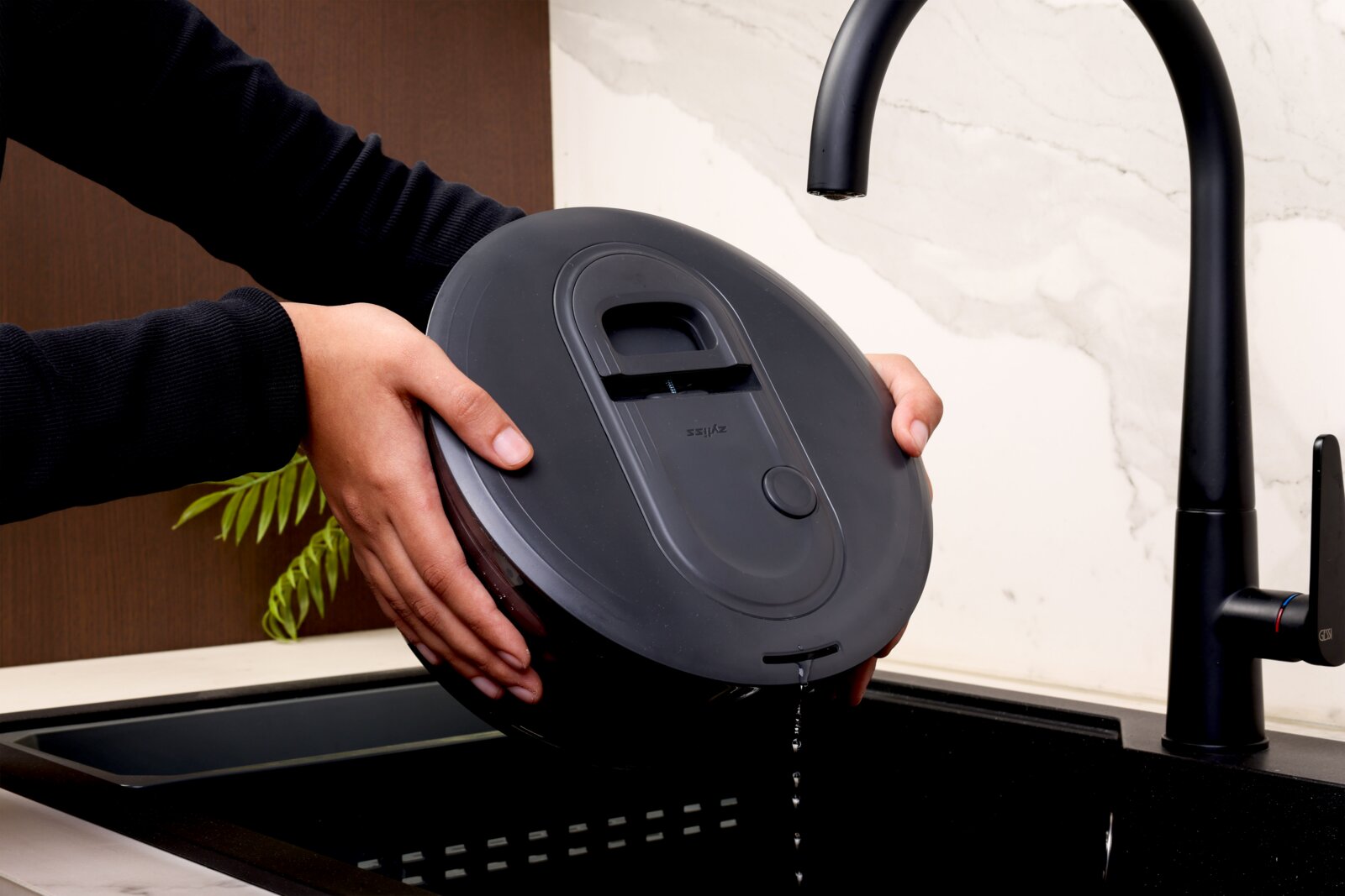 Schuine zijaanzicht van een persoon die een zwarte elektrische rijstkoker/ multicooker boven een gootsteen kantelt om water af te gieten, met een zwarte keukenkraan op de achtergrond.