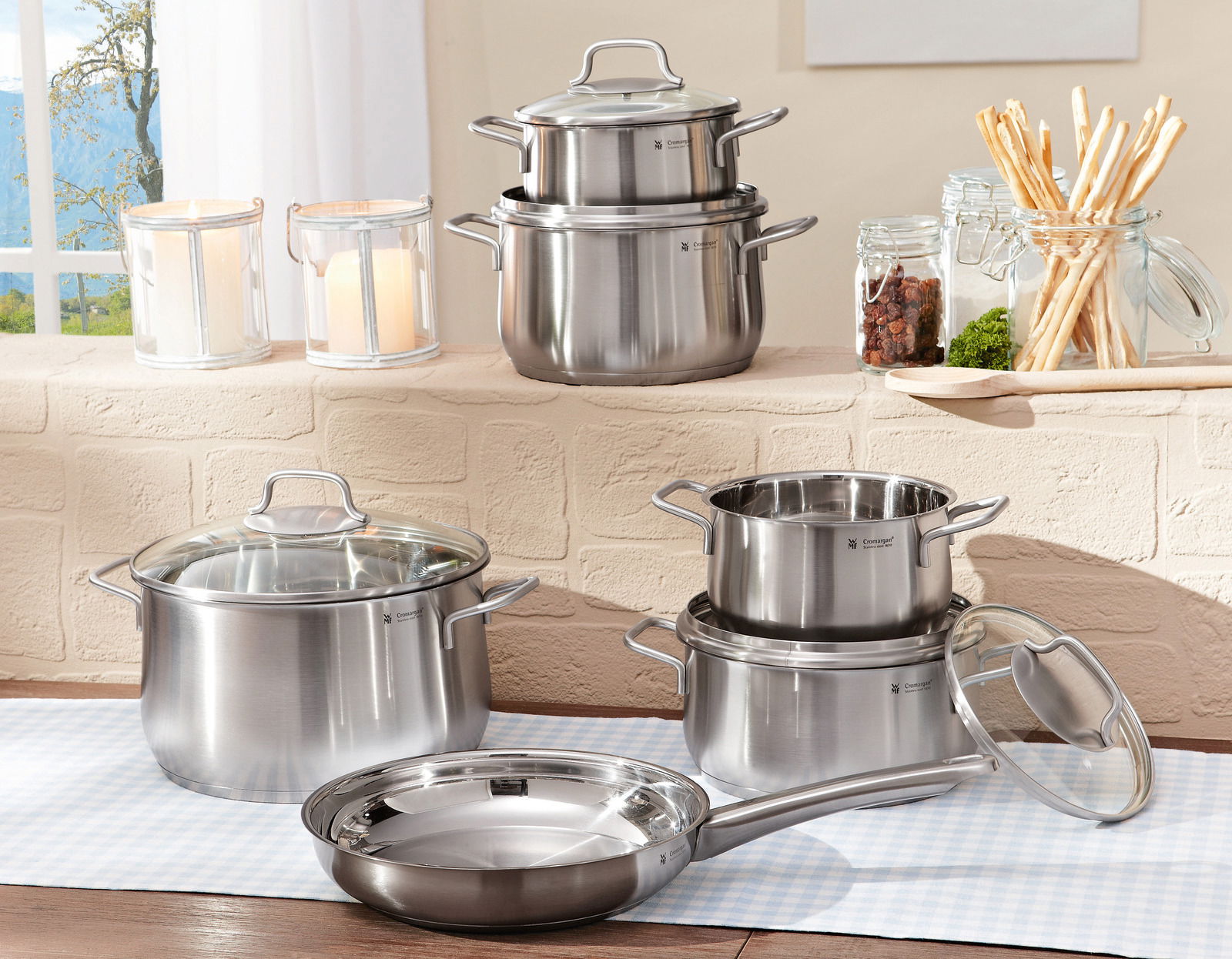 Topfset Brillant 6-tlg. aus Edelstahl, bestehend aus mehreren Töpfen und einer Pfanne mit Glasdeckeln, auf einem Tisch in einer hellen Küche präsentiert. Perspektive: seitlich.