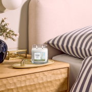 Brandende Yankee Candle Clean Cotton geurkaars met twee lonten in een glazen pot, vanuit schuin vooraanzicht op een houten nachtkastje naast een bed
