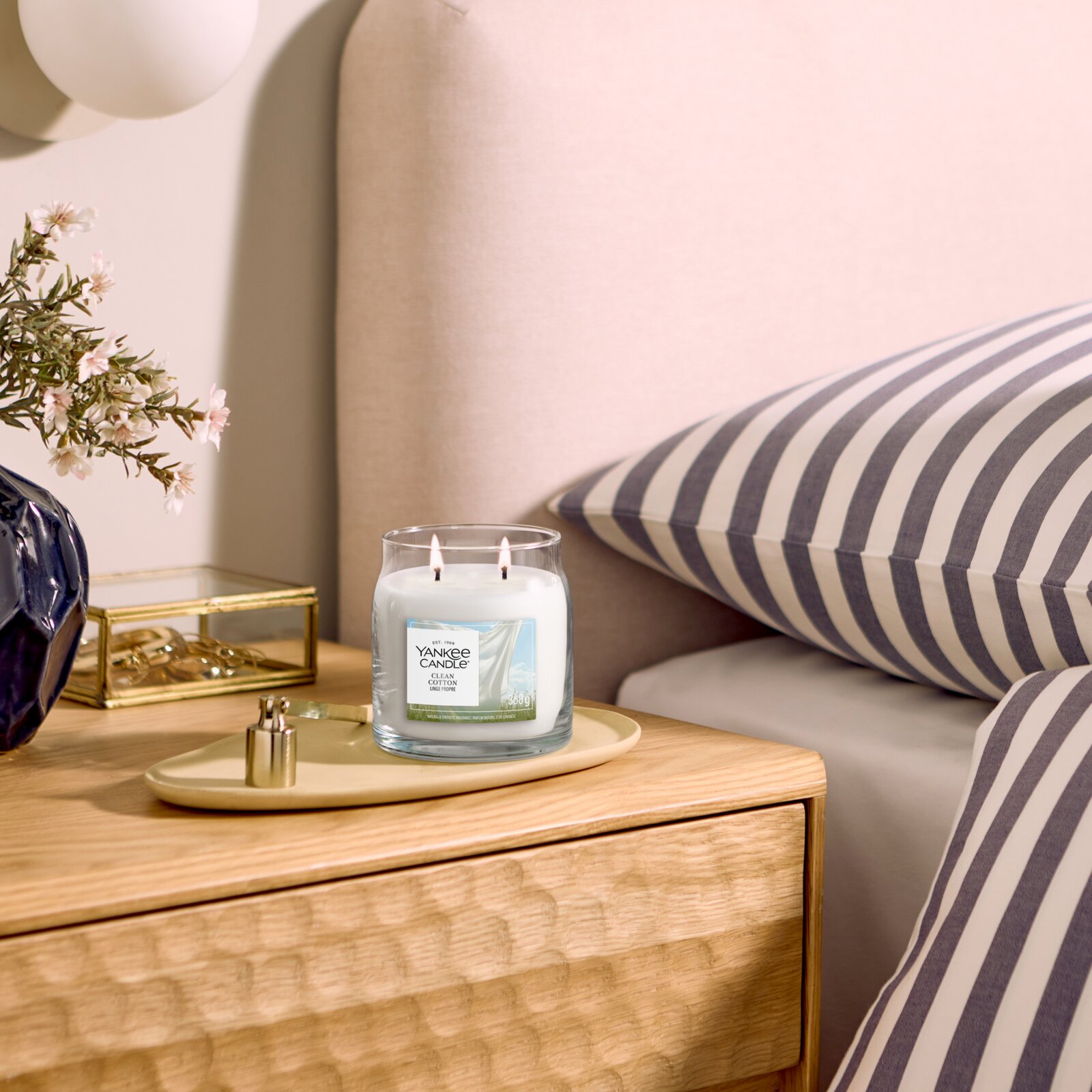 Brandende Yankee Candle Clean Cotton geurkaars met twee lonten in een glazen pot, vanuit schuin vooraanzicht op een houten nachtkastje naast een bed