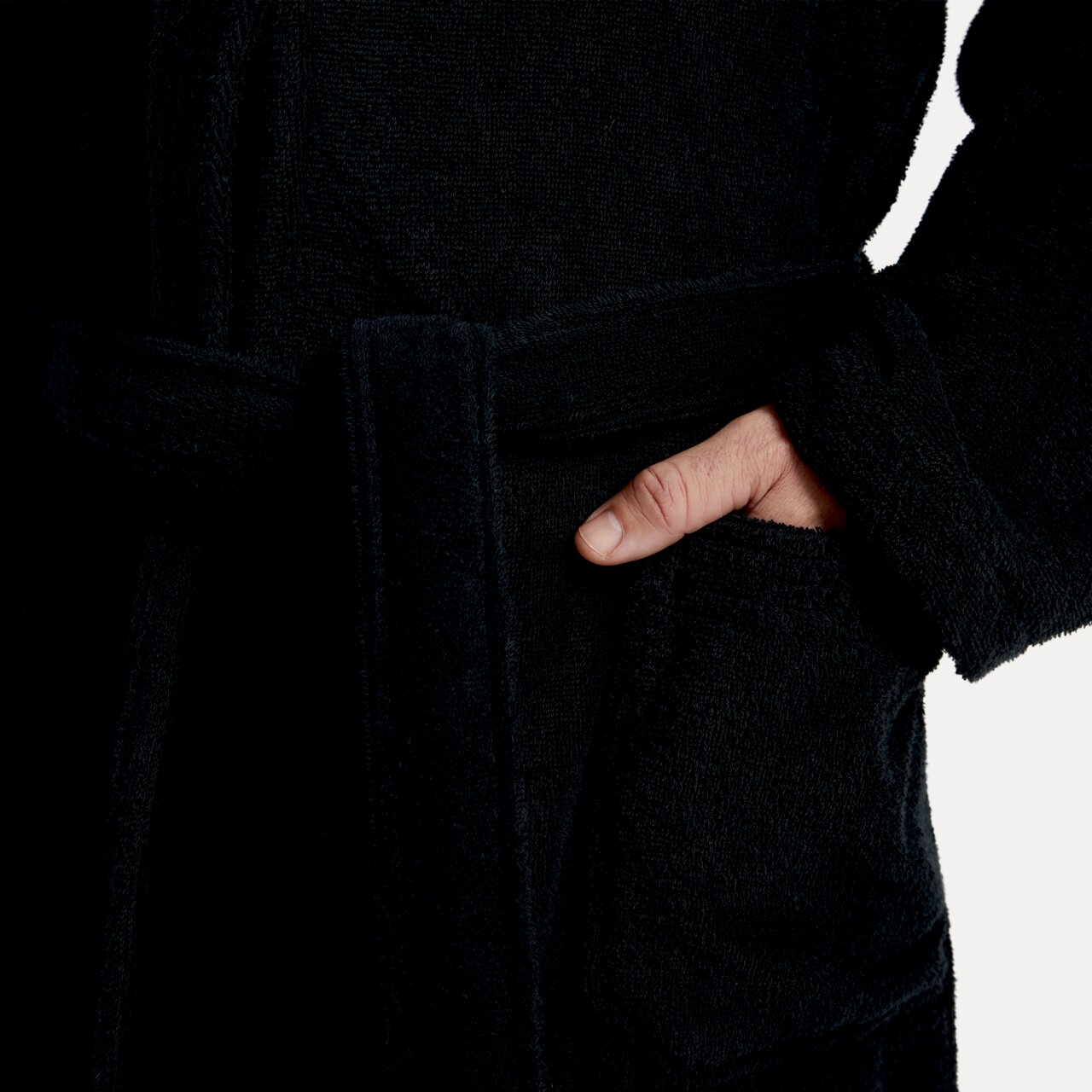 Close-up van een zwarte badjas met sjaalkraag, ceintuur en zak, met een hand in de zak.