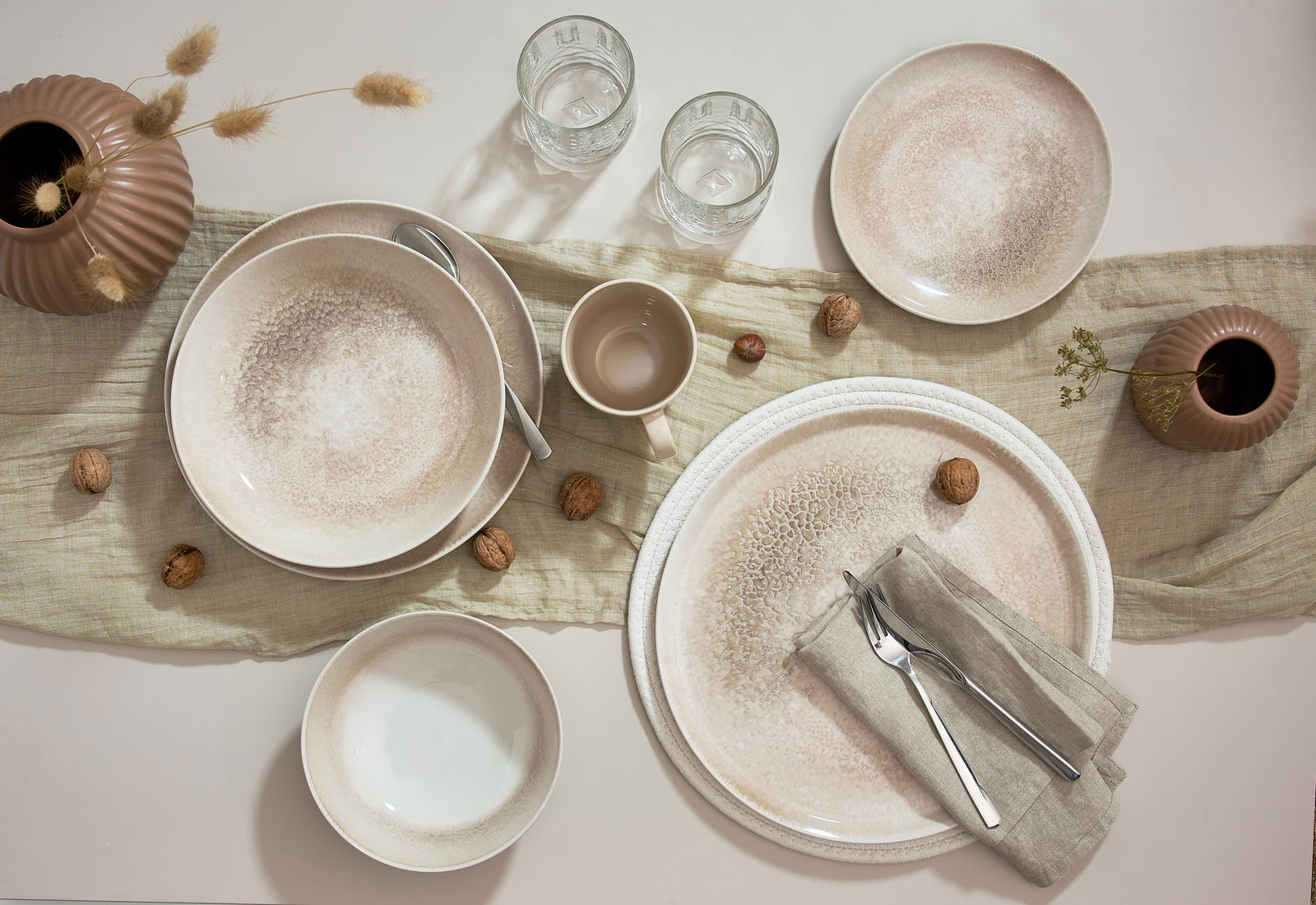 Vogelperspektive auf ein 18-teiliges Tellerset mit natürlichem Design, bestehend aus Tellern, Schalen und Tassen, arrangiert auf einem Tisch mit Leinentuch, Gläsern und Dekoration.