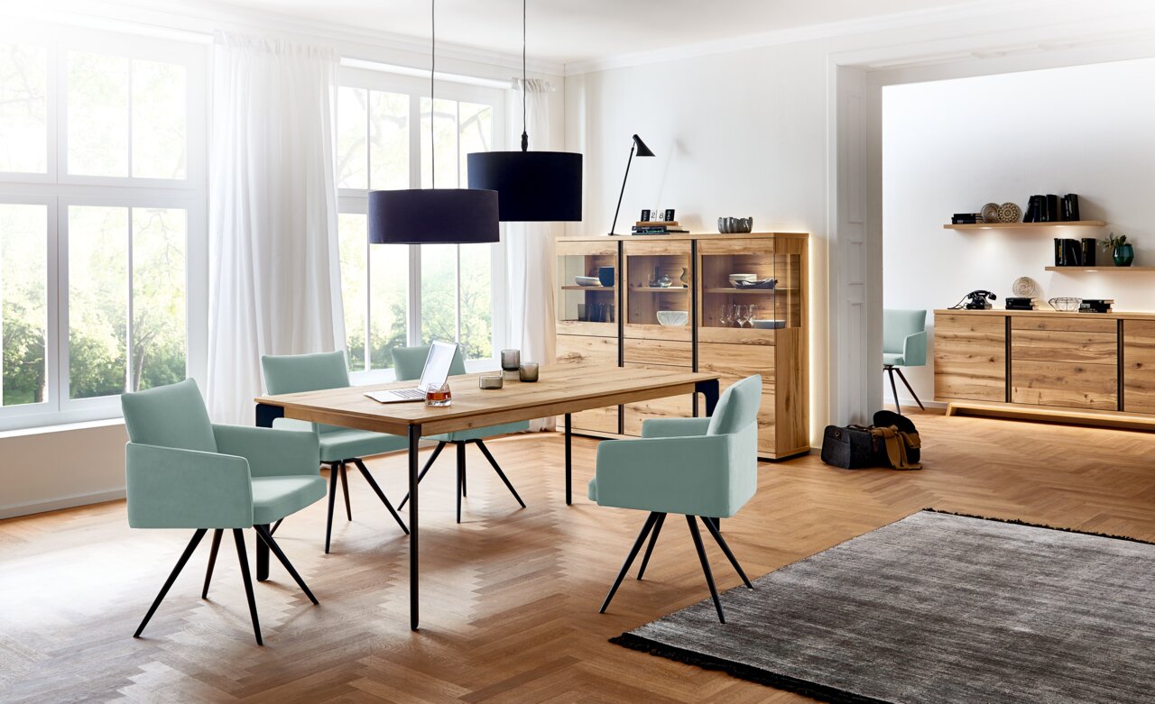 Eetkamer met een moderne houten tafel, omringd door mintgroene stoelen, op de voorgrond. Op de achtergrond een hoog kastmeubel met glazen vitrine en ledverlichting, en een dressoir tegen de muur. Perspectief: zijaanzicht van de eetkamer met uitzicht op he