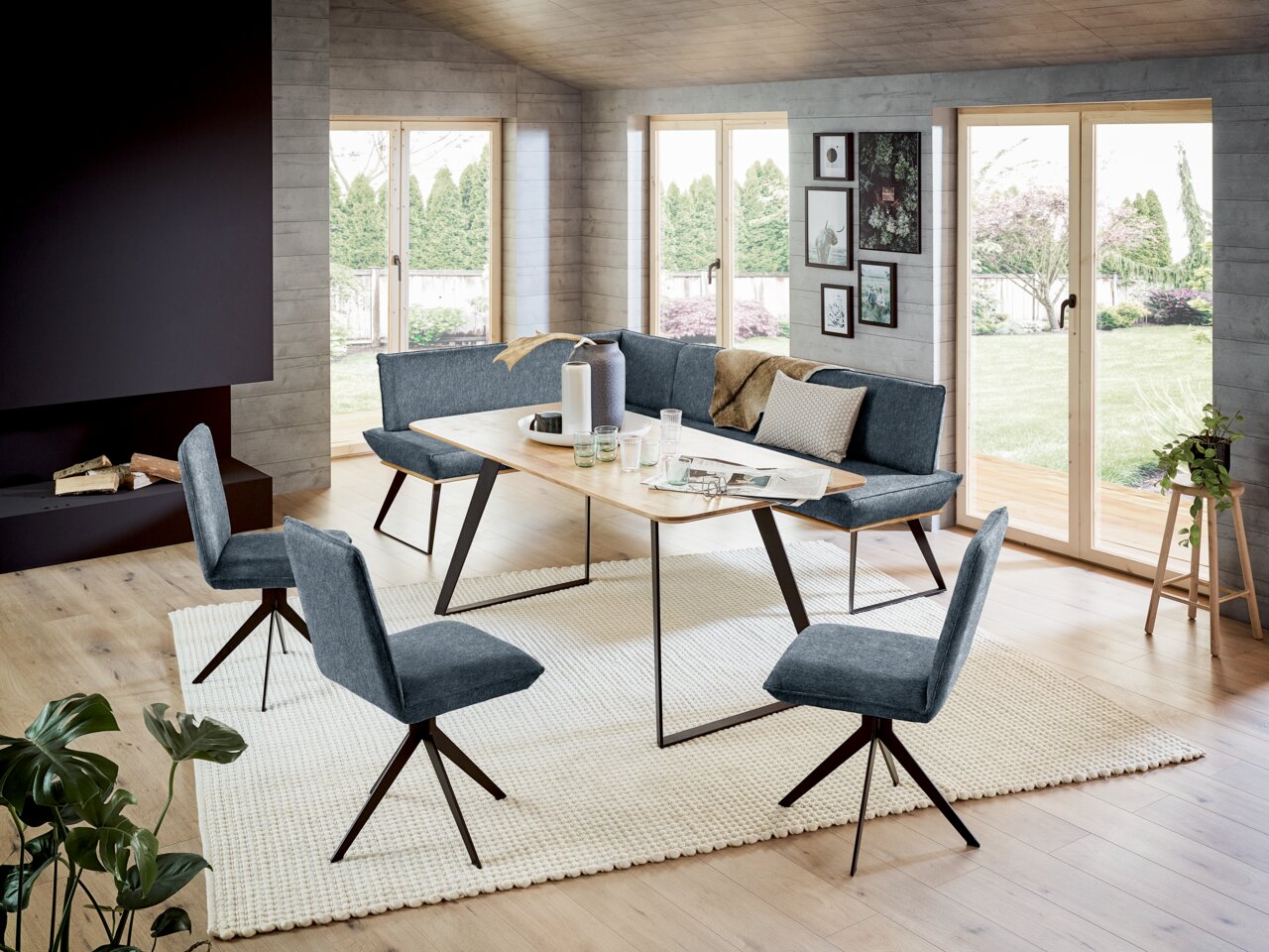 Moderne Eckbankgruppe in einem hellen Wohnzimmer mit großen Fenstern, bestehend aus einer Eckbank und vier Stühlen in grauem Stoff, sowie einem Holztisch mit Metallbeinen. Perspektive von schräg vorne.