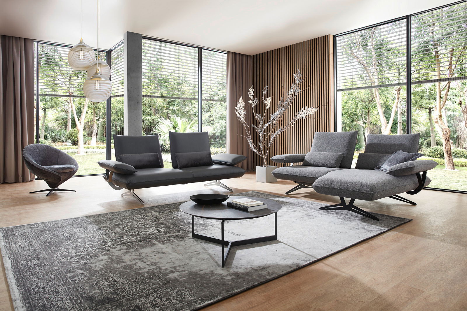 Modernes Wohnzimmer mit großen Fenstern, einem grauen Sessel und Sofa-Set, einem runden Couchtisch und einem eleganten Teppich, seitliche Perspektive.