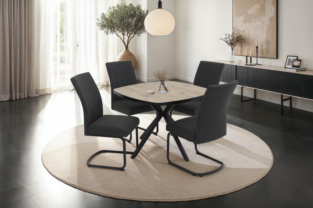 Z2 Esstisch ROTTERDAM Moderner Esstisch mit runder Holzplatte und schwarzen Metallbeinen, umgeben von vier dunkel gepolsterten Stühlen auf einem hellen Teppich. Perspektive: seitlich.