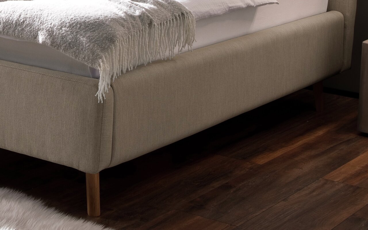 Nahaufnahme eines Polsterbetts in Beige mit Holzfüßen, seitliche Perspektive