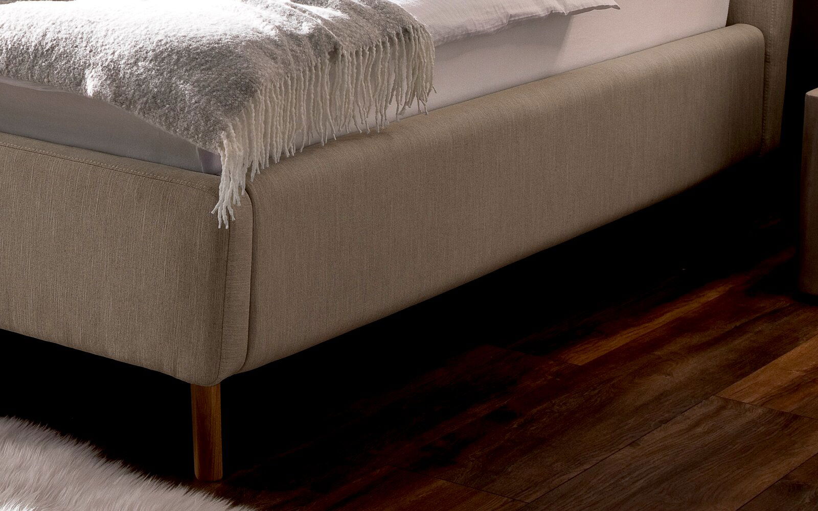 Nahaufnahme eines Polsterbetts in Beige mit Holzfüßen, seitliche Perspektive