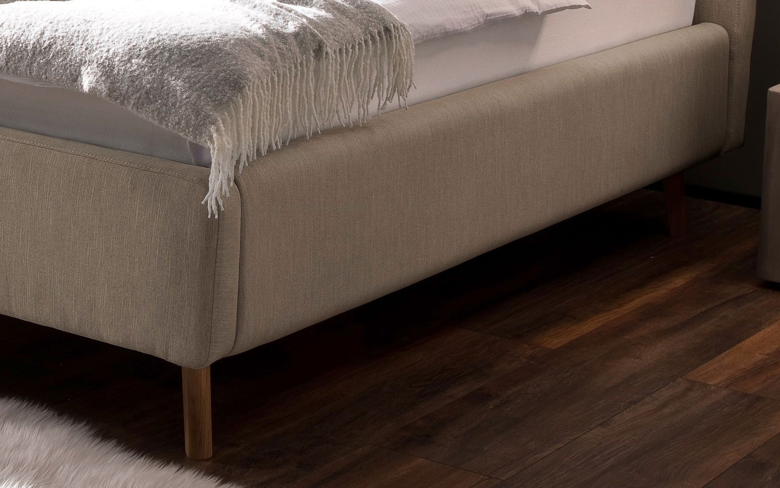 Nahaufnahme eines Polsterbetts in Beige mit Holzfüßen, seitliche Perspektive