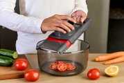 Person benutzt einen grauen Gemüsehobel/Mandoline mit Handschutz über einer transparenten Schüssel, Tomatenscheiben fallen hinein; seitliche Perspektive in der Küche mit Gurken, Tomaten, Karotten und halber Zitrone auf dem Tisch.
