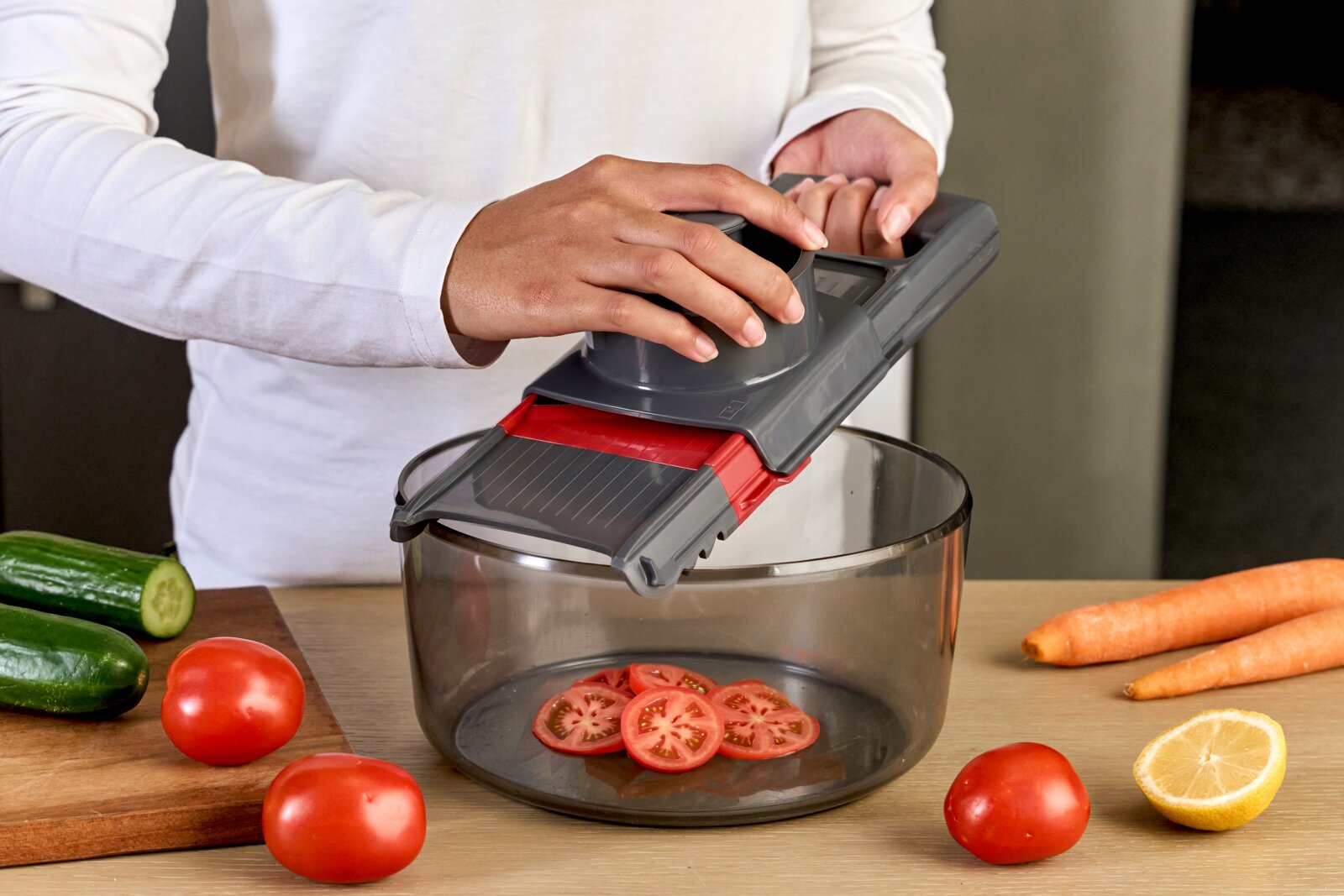 Person benutzt einen grauen Gemüsehobel/Mandoline mit Handschutz über einer transparenten Schüssel, Tomatenscheiben fallen hinein; seitliche Perspektive in der Küche mit Gurken, Tomaten, Karotten und halber Zitrone auf dem Tisch.