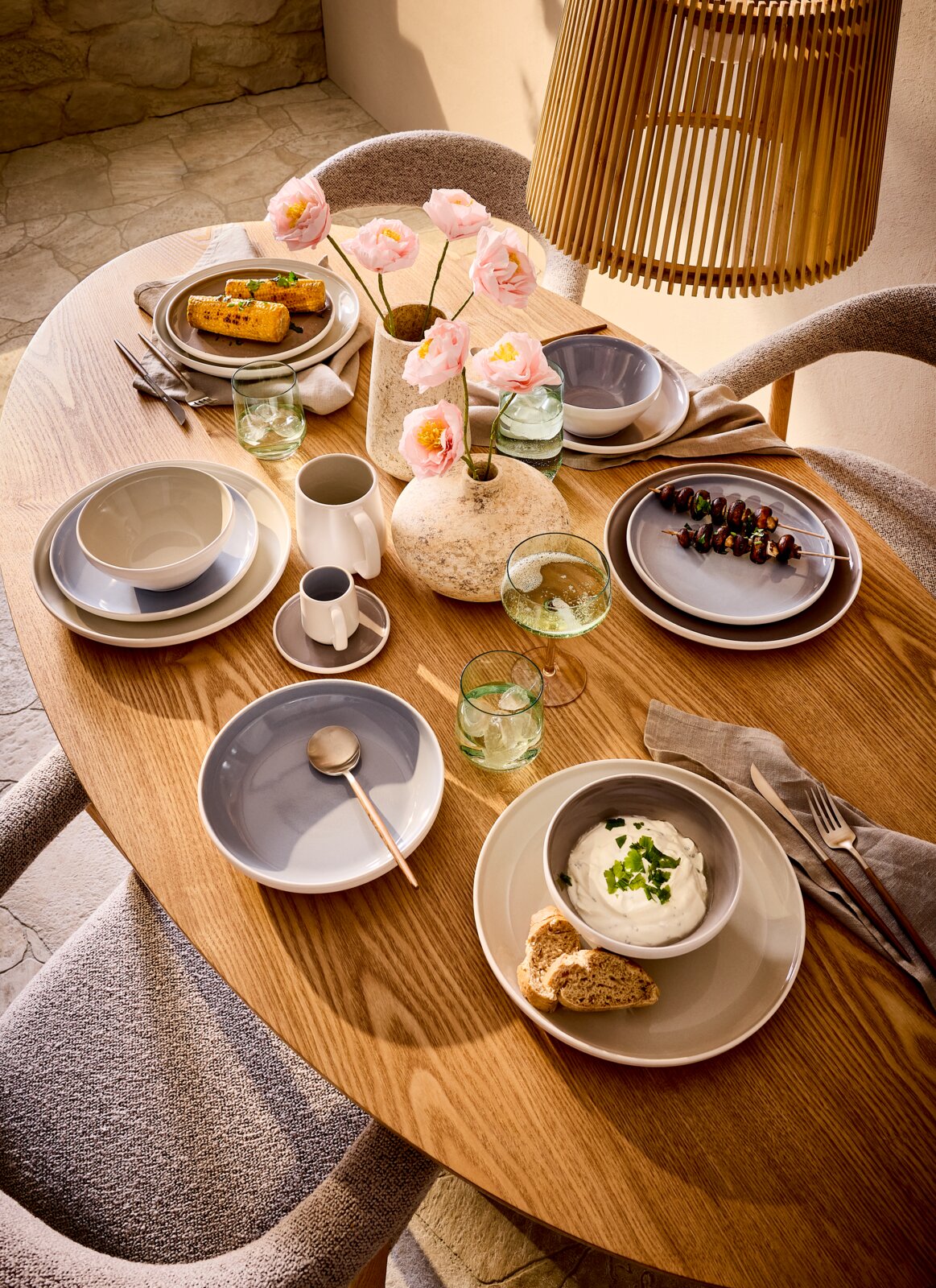 Novel Dessertteller LEVANTO Schräg von oben fotografierter, ovaler Holzesstisch mit modernem graubeigem Geschirr, Gläsern, Besteck, zwei Natursteinvasen mit rosa Blumen und dekorativ angerichteten Speisen in warmem Sonnenlicht.