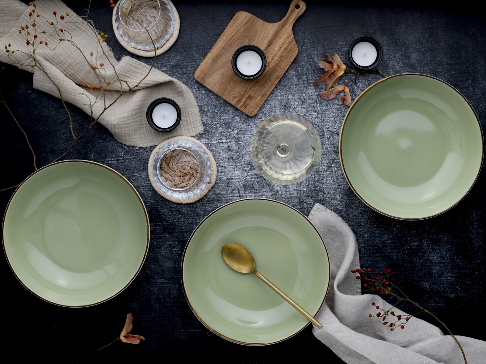 Draufsicht auf ein stilvolles Tisch-Arrangement mit drei großen, hellgrünen Keramikschalen, goldfarbenem Löffel, Teelichtern, Holzbrett, Leinentüchern und dekorativen Zweigen auf dunkler Oberfläche.