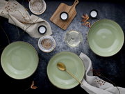 Draufsicht auf ein stilvolles Tisch-Arrangement mit drei großen, hellgrünen Keramikschalen, goldfarbenem Löffel, Teelichtern, Holzbrett, Leinentüchern und dekorativen Zweigen auf dunkler Oberfläche.
