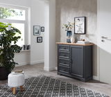 Dunkelgraue Kommode mit vier Schubladen und einer Tür, dekoriert mit Vasen und Büchern, in einem modernen, hellen Raum mit Teppich und Pflanzen, seitliche Perspektive.