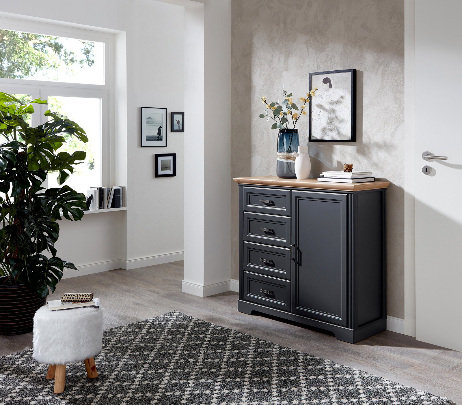 Landscape Kommode JUDITH Dunkelgraue Kommode mit vier Schubladen und einer Tür, dekoriert mit Vasen und Büchern, in einem modernen, hellen Raum mit Teppich und Pflanzen, seitliche Perspektive.