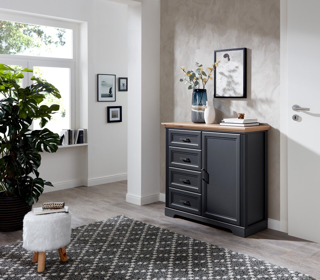 Landscape Kommode JUDITH Dunkelgraue Kommode mit vier Schubladen und einer Tür, dekoriert mit Vasen und Büchern, in einem modernen, hellen Raum mit Teppich und Pflanzen, seitliche Perspektive.