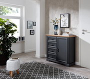 Dunkelgraue Kommode mit vier Schubladen und einer Tür, dekoriert mit Vasen und Büchern, in einem modernen, hellen Raum mit Teppich und Pflanzen, seitliche Perspektive.