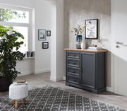 Dunkelgraue Kommode mit vier Schubladen und einer Tür, dekoriert mit Vasen und Büchern, in einem modernen, hellen Raum mit Teppich und Pflanzen, seitliche Perspektive.