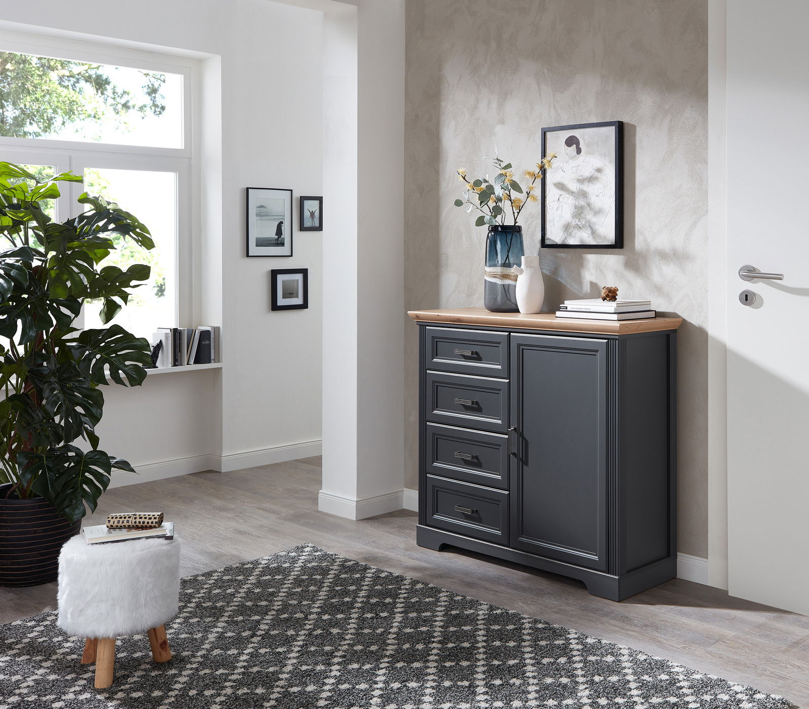 Dunkelgraue Kommode mit vier Schubladen und einer Tür, dekoriert mit Vasen und Büchern, in einem modernen, hellen Raum mit Teppich und Pflanzen, seitliche Perspektive.