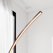 Moderne, schlanke LED-Bogen-Stehleuchte mit warmweiß leuchtendem Lichtband in schwarzem Rahmen, Detailaufnahme aus seitlicher Perspektive vor hellem Wohnraum-Hintergrund