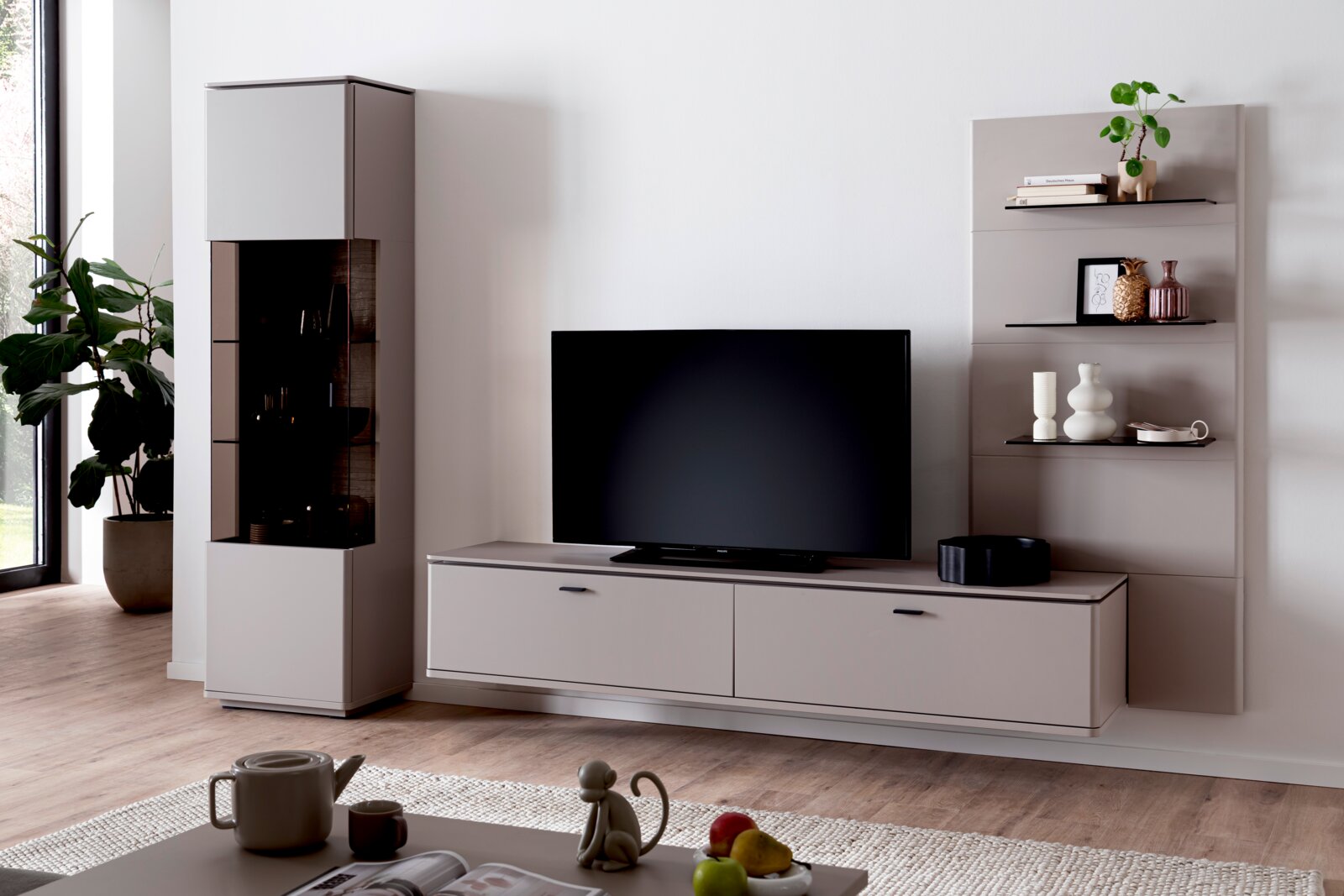 Zurbrüggen Wandkombination TORRENT Frontale Wohnzimmeransicht: modernes TV-Lowboard in Hellgrau mit Flachbildfernseher, links eine hohe Vitrine mit Glasfront und rechts ein Regal mit Dekoration vor weißer Wand.
