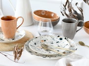 Stilvolles Geschirr-Arrangement mit gesprenkelten Keramiktellern, goldfarbenem Besteck, grauer und pfirsichfarbener Tasse, Schalen und Glas auf weißem Tisch, fotografiert in leicht erhöhter Frontperspektive.