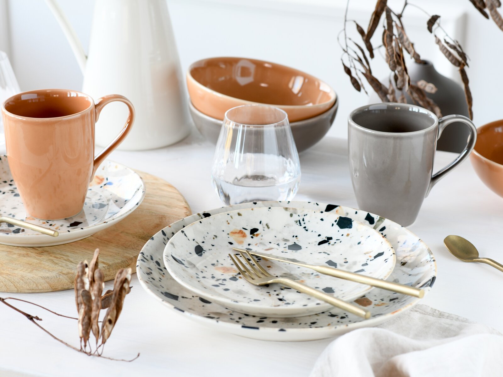 Stilvolles Geschirr-Arrangement mit gesprenkelten Keramiktellern, goldfarbenem Besteck, grauer und pfirsichfarbener Tasse, Schalen und Glas auf weißem Tisch, fotografiert in leicht erhöhter Frontperspektive.