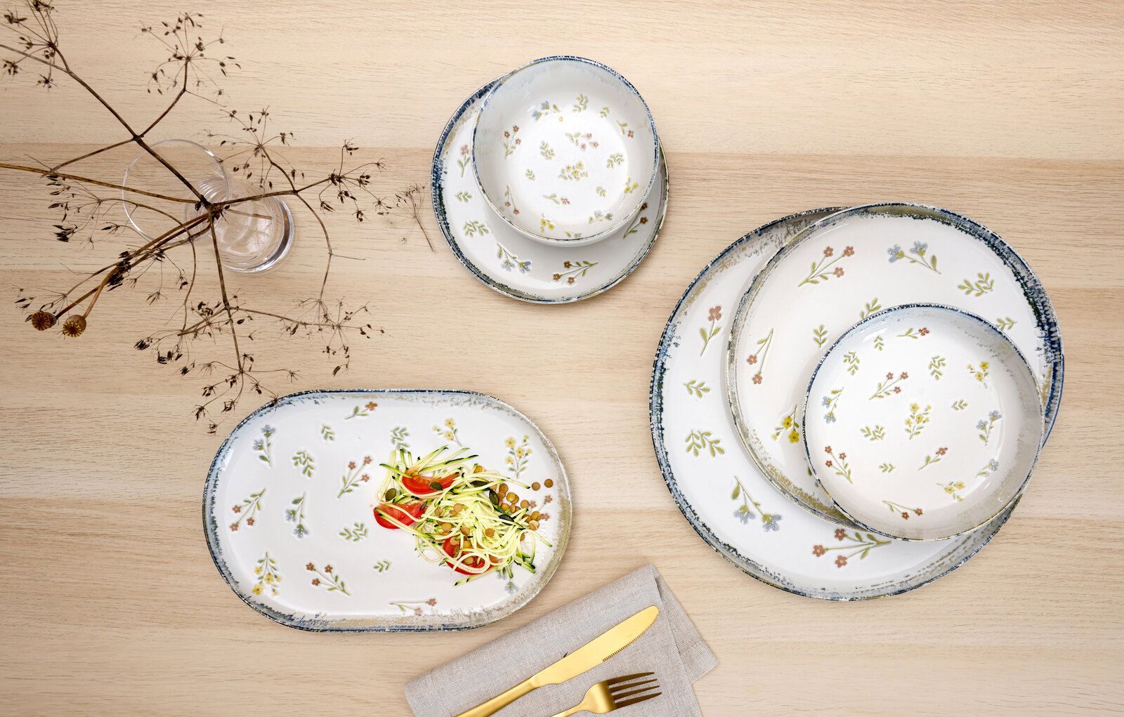 Draufsicht auf ein weißes Keramik-Geschirrset mit zartem Blumenmuster und blau gesprenkeltem Rand: ovaler Servierteller mit Salat, zwei runde Teller und eine Schale auf hellem Holztisch, daneben Glasvase mit Trockenblumen und Besteck auf Serviette.
