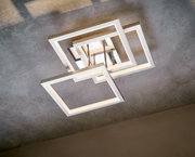 Moderne led-plafondlamp met overlappende vierkante frames van geborsteld metaal, met een warmwitte lichtkleur, gemonteerd op een betonnen plafond, gezien van onderaf in een licht schuin perspectief.