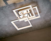 Moderne LED-Deckenleuchte mit überlappenden quadratischen Rahmen in gebürstetem Metall, warmweiß leuchtend, an Betondecke montiert, Ansicht von unten in leichter Schrägperspektive.