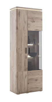 Hohe Vitrine aus heller Eiche-Optik mit Glasvitrine und LED-Beleuchtung, schwarze Griffe, freigestellt vor weißem Hintergrund, Ansicht schräg von vorn links