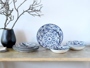 Blau-weiß gemustertes Keramik-Geschirrset mit Tellern und Schalen auf einem Holztisch, links schwarze Vase mit Zweigen, frontal aus leicht erhöhter Perspektive fotografiert