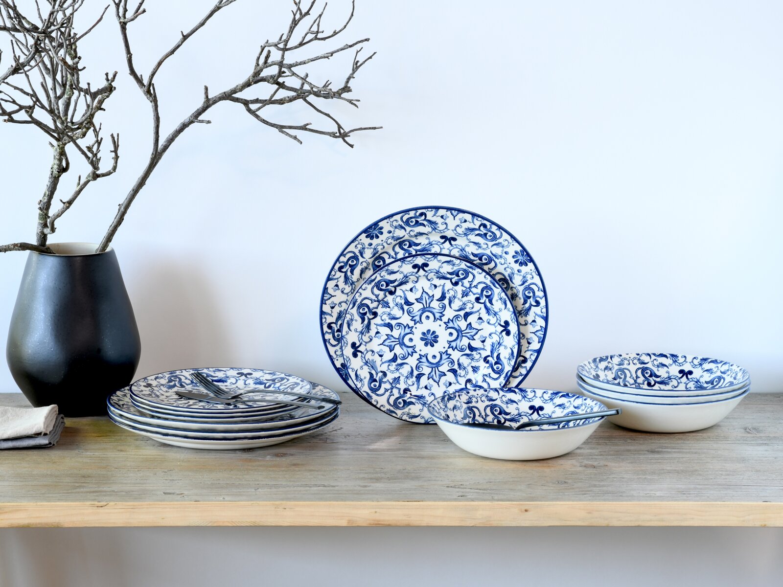 Blau-weiß gemustertes Keramik-Geschirrset mit Tellern und Schalen auf einem Holztisch, links schwarze Vase mit Zweigen, frontal aus leicht erhöhter Perspektive fotografiert