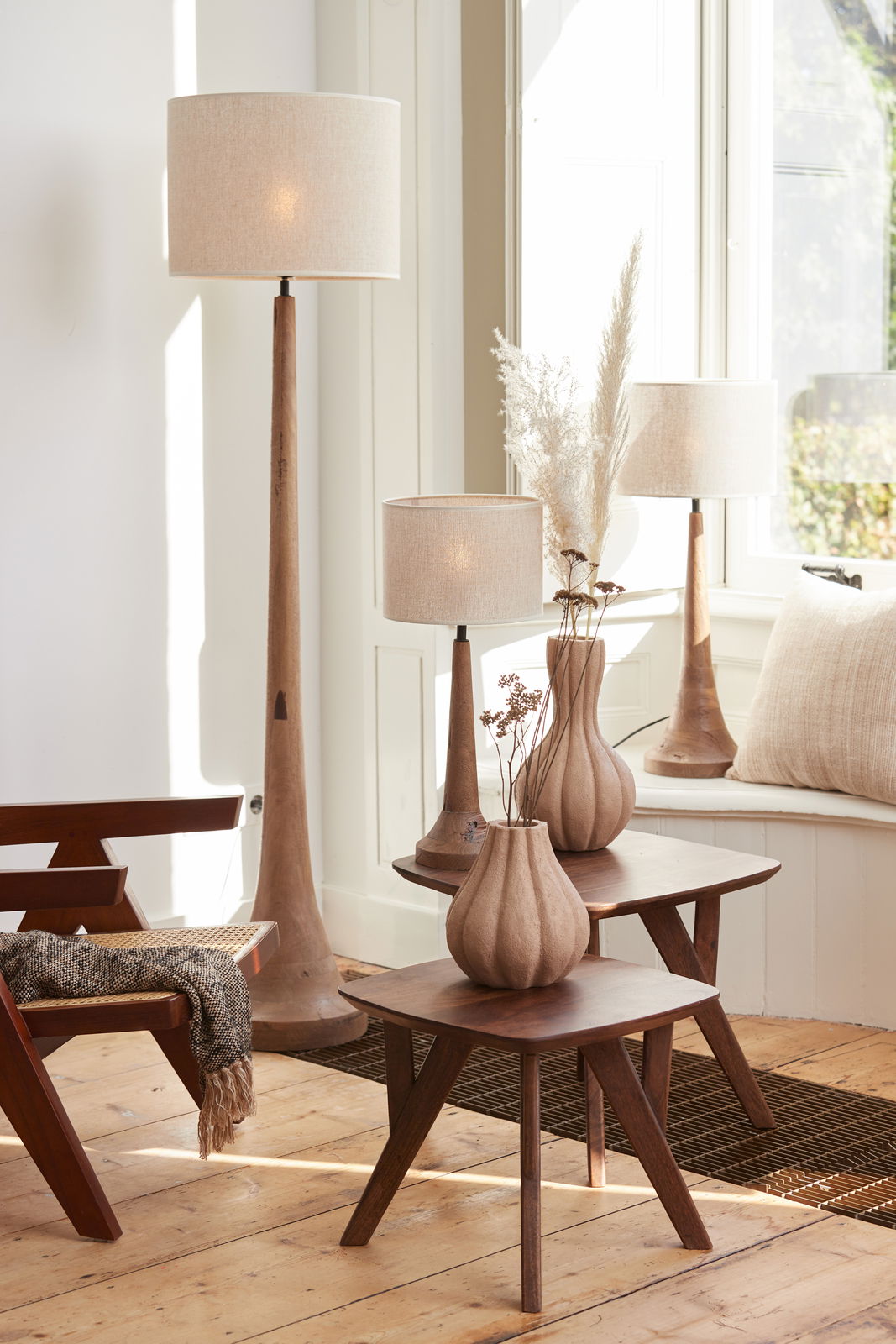Ein stilvoller Raum mit mehreren Lampen mit beigen Lampenschirmen, die auf einem Holztisch und einer Fensterbank stehen. Die Perspektive zeigt die Lampen von der Seite, mit natürlichem Licht, das durch das Fenster fällt.