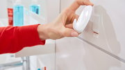 Nahaufnahme einer Hand, die einen weißen tesa Powerstrips Haken Large an einer Wand im Badezimmer anbringt. Perspektive von der Seite.