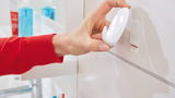 Nahaufnahme einer Hand, die einen weißen tesa Powerstrips Haken Large an einer Wand im Badezimmer anbringt. Perspektive von der Seite.