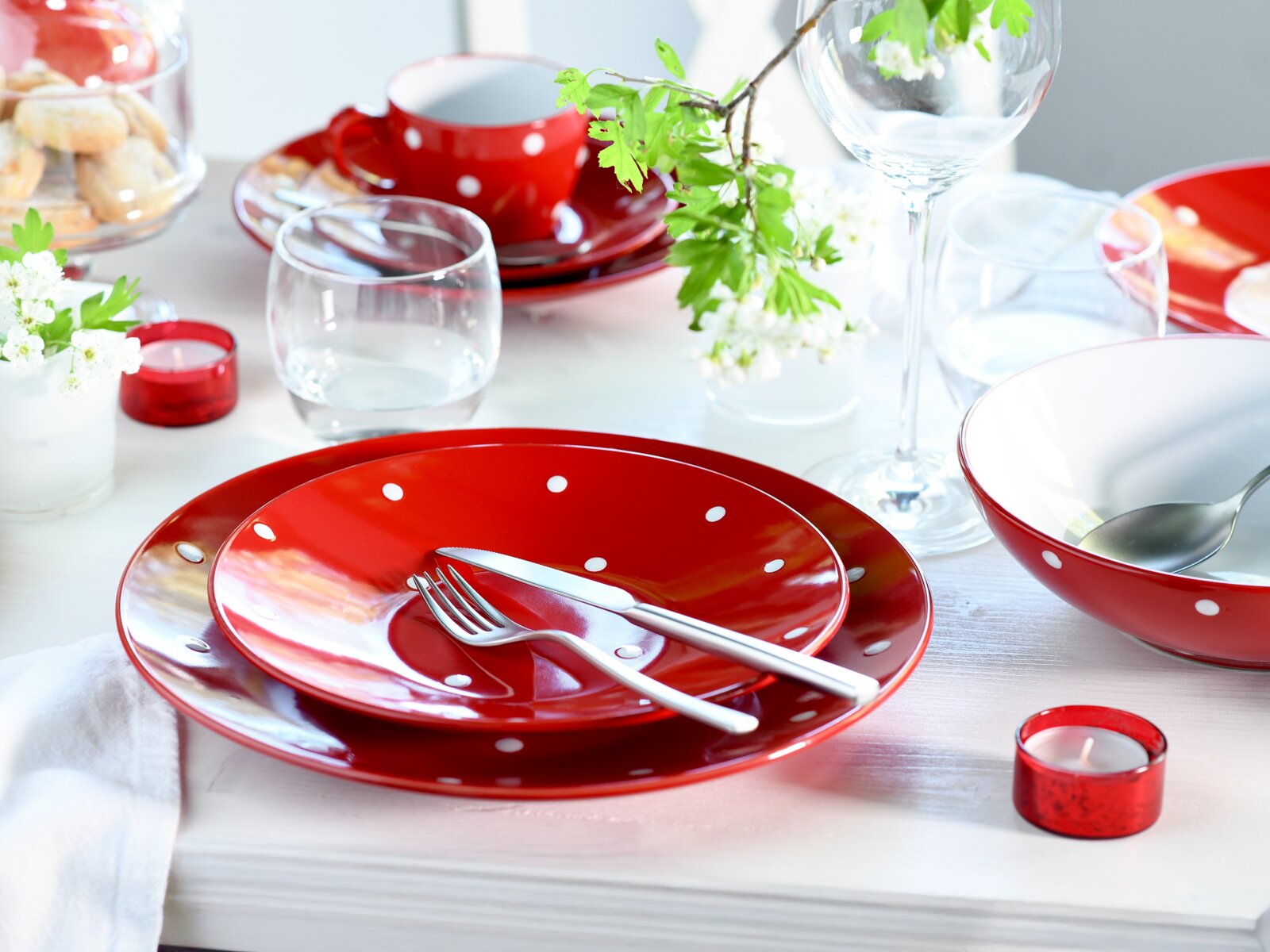 Schwarz-weiß gepunktetes, rotes Keramikgeschirr-Set (Teller, Schale und Tasse) auf weiß gedecktem Tisch mit Besteck und Gläsern, fotografiert in schräger Draufsicht von vorn.