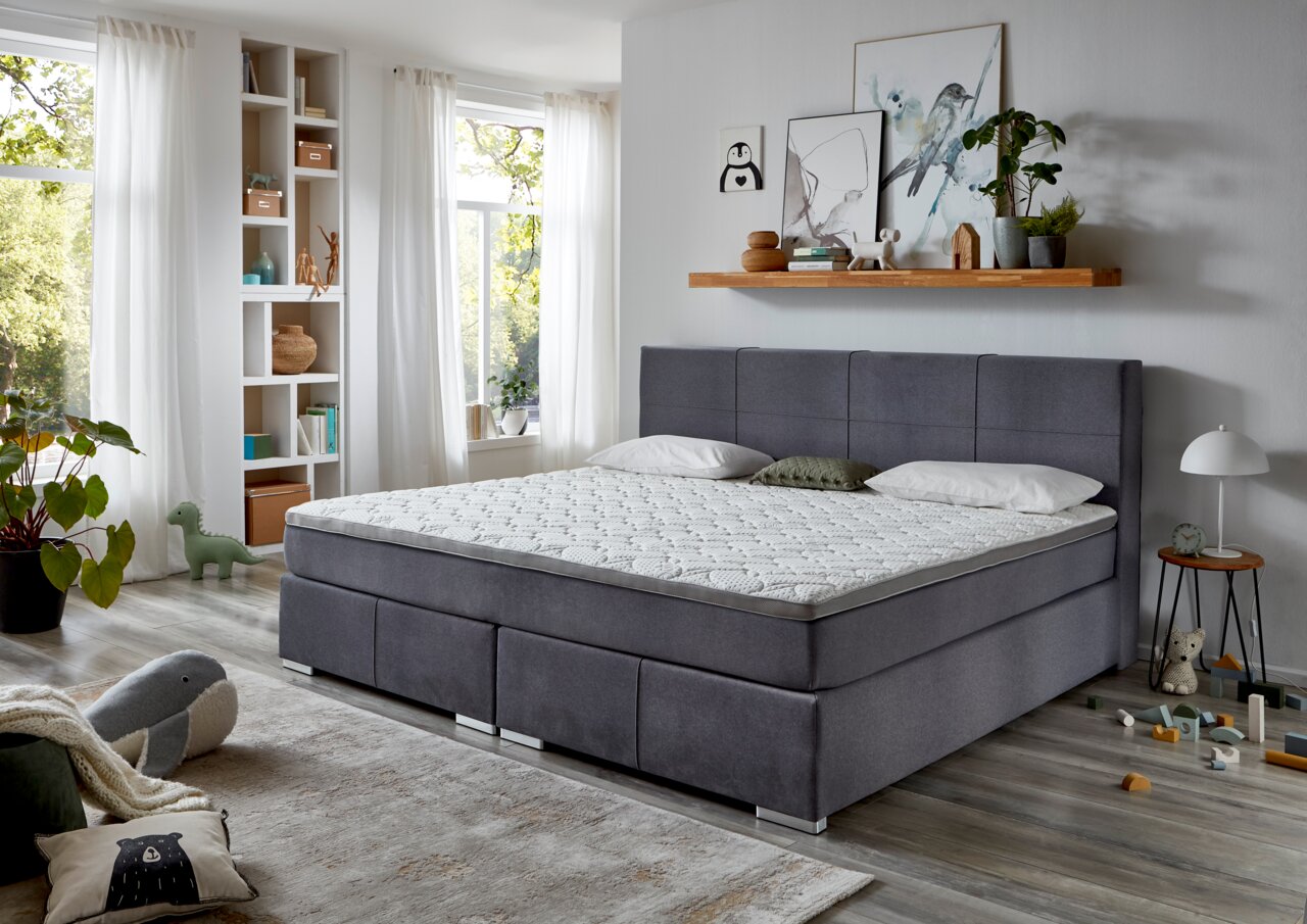 Graues Boxspringbett mit hohem gepolstertem Kopfteil, weißem Topper und Kissen in einem hellen, modern eingerichteten Schlafzimmer, aus schräger Frontansicht.