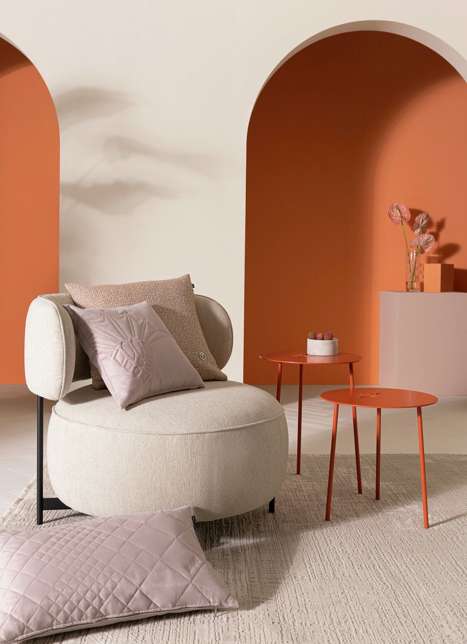 Gemütliche Wohnzimmerszene mit einem beigen Sessel, dekoriert mit zwei Zierkissen in verschiedenen Texturen und Farben, vor einem modernen Hintergrund in warmen Erdtönen. Perspektive: Frontalansicht.