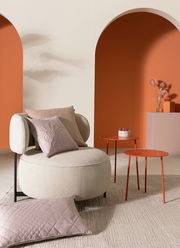 Gemütliche Wohnzimmerszene mit einem beigen Sessel, dekoriert mit zwei Zierkissen in verschiedenen Texturen und Farben, vor einem modernen Hintergrund in warmen Erdtönen. Perspektive: Frontalansicht.